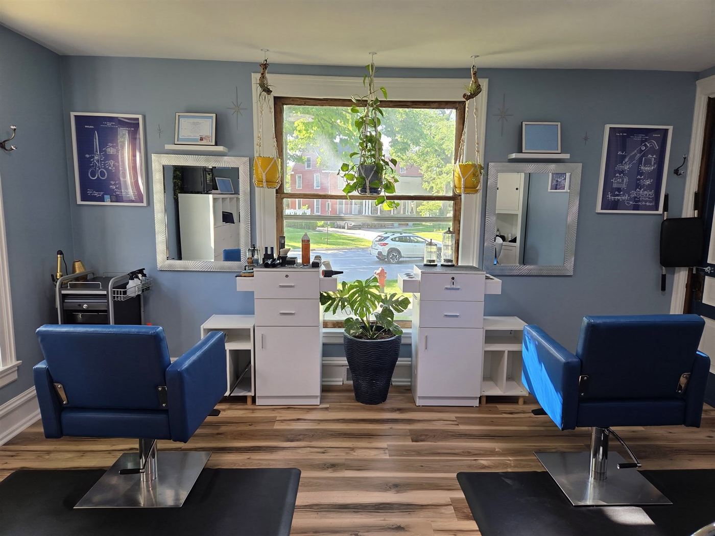 Inside Blueprint Hair Designs: two blue styling chairs, a sunny front window with hanging plants, and framed blueprint-style patent drawings of scissors and clippers